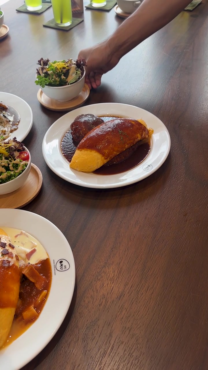 Una mano posiziona un'insalatiera accanto a omelette di riso, zuppa e un piatto servito in un luminoso ambiente da caffè.