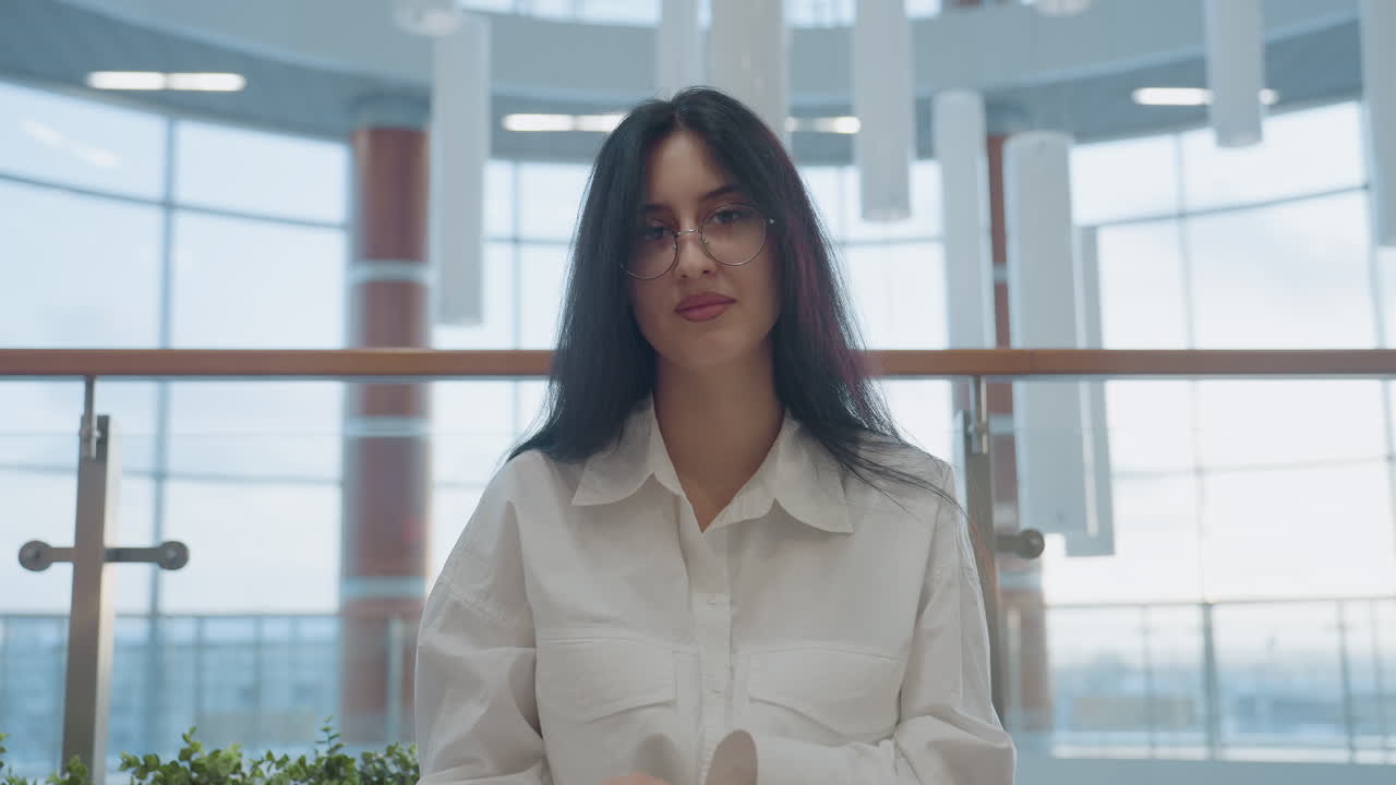 Close up of young woman seated on modern bench gently closes book in her hand, surrounded by plants and contemporary glass interior, expressing calm mood and quiet focus in spacious setting