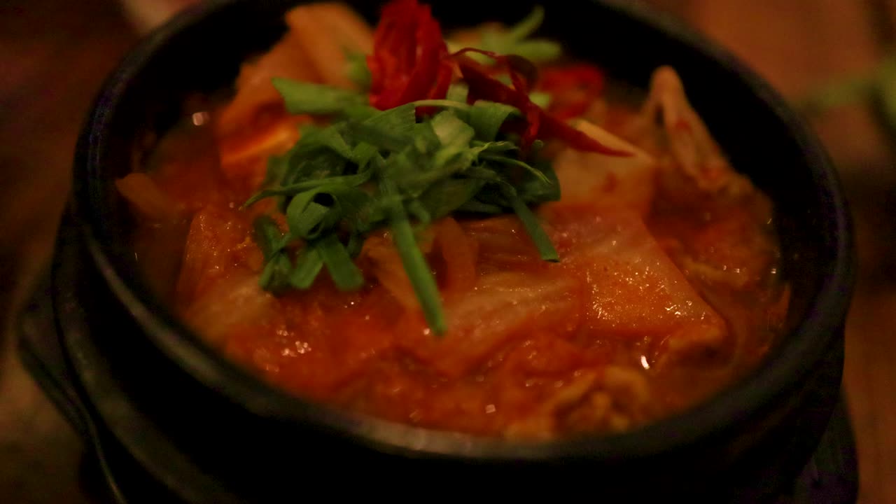 primer plano de sopa de kimchi burbujeante en un cuenco de piedra, adornada con verduras frescas, bajo una iluminación ambiente cálida
