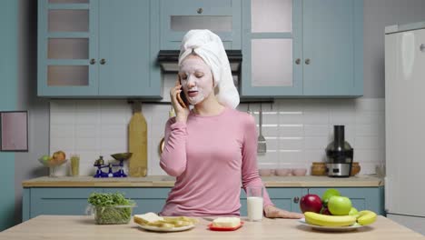 woman talking on phone while wearing a face mask and having breakfast in the kitchen