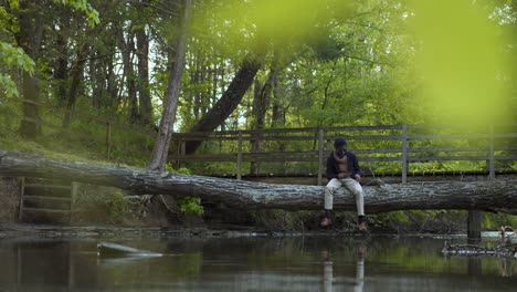 man in the woods seated on a tree trunk bridge reflecting over water