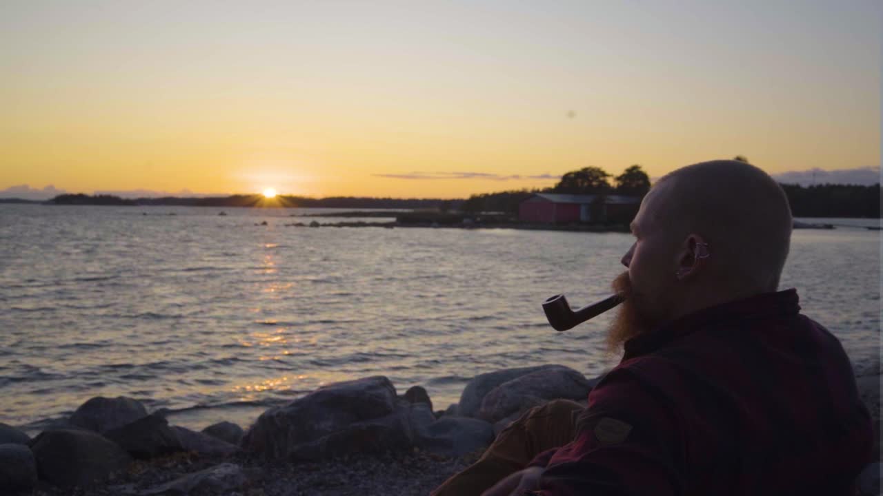 hombre sigma disfrutando de un tiempo de calidad pacífico por sí mismo mientras fuma soplando una pipa antigua de época sosteniendo en su boca mientras el sol se pone produciendo rayas rayos de luz naturaleza clamness