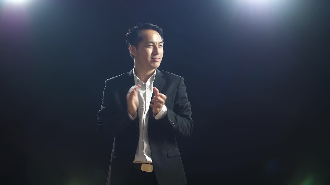 블랙 스크린 스튜디오에 서 있는 동안 손을 박고 주변을 둘러보는 미소 짓는 아시아 스피커 남자