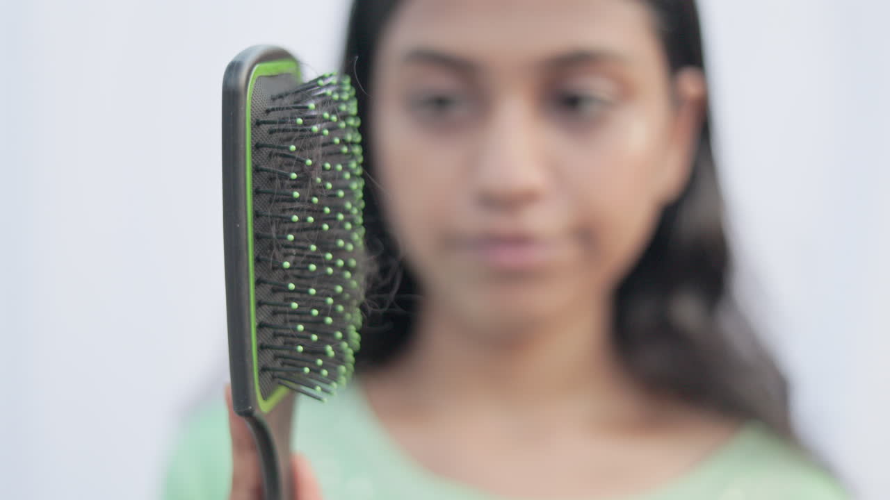 Indian young woman removing lost hair from the comb and get shocked in white background, focus on foreground.