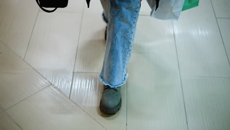 leg view of individual in jeans and boots carrying shopping bag in left hand and black bag in right, walking in mall, shadow on tile floor