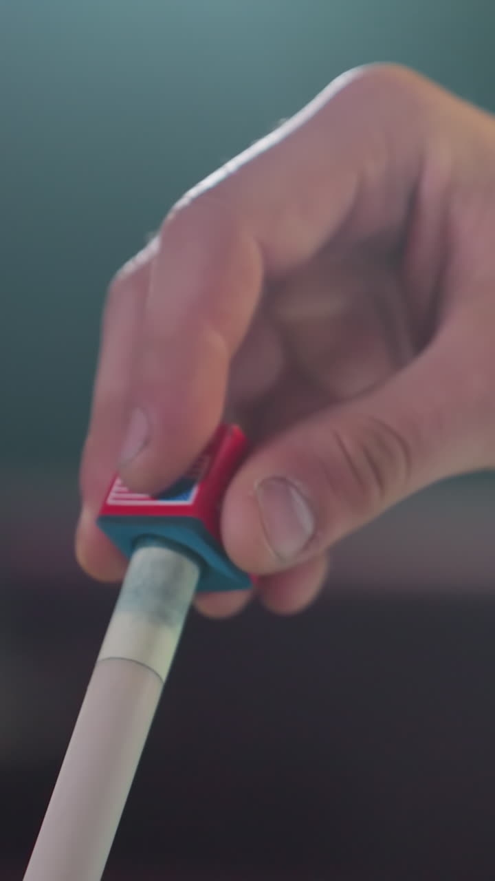Primo piano di una mano in una camicia bianca che applica gesso rosso e blu a una stecca da biliardo prima di una partita. La presa decisa e lo sfondo sfocato enfatizzano la preparazione per la prossima mossa.