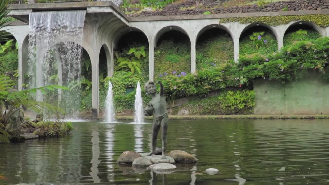 Aerial drone shot showing lush pond garden with bronze statue and cascading water under stone arches surrounded by greenery and blooming plants creating scene at Monte Palace Tropical Garden