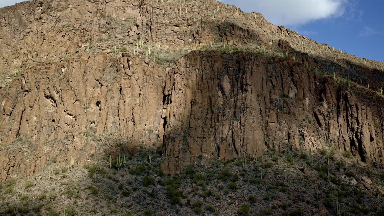 드론은 사막의 산을 향해 날아가며, 티로 여 있으며, 구름의 그림자가 풍경을 가로질러 움직이고 있으며, 배경에는 큰 은 바위 산이 있습니다.