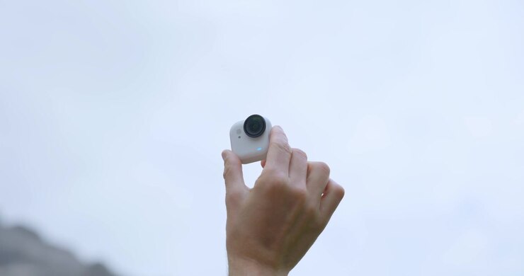 Close-up da mão de uma pessoa segurando uma pequena câmera Insta360 GO branca ao ar livre com céu desfocado ao fundo. Espaço para texto.