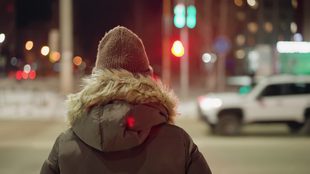 View of a woman in a winter coat with a fur hood, walking across a road at night with a green and red traffic signal and blurry lights in the background. A white car is moving in the distance