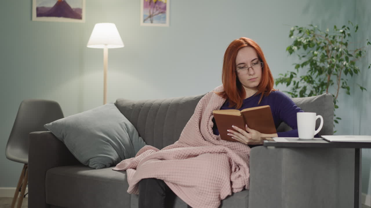 Redhead woman reads interesting book sitting on sofa