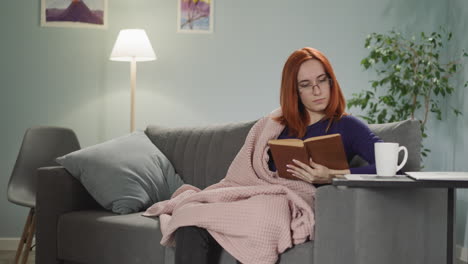 redhead woman reads interesting book sitting on sofa