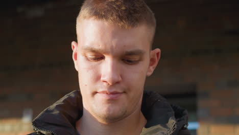 portrait view of fair skinned man standing outdoors with head slightly bowed in deep reflection, wearing black padded vest and neutral expression against blurred brick wall background