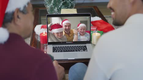 Padre-E-Hijo-Birraciales-Sonrientes-Usando-Una-Computadora-Portátil-Para-Una-Videollamada-Navideña-Con-La-Familia-En-La-Pantalla