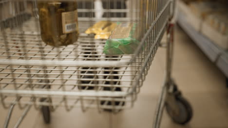 close up shot of hand placing glass jar of pickled cucumbers into metal shopping cart grid alongside bars and vegetables on lower rack during busy supermarket grocery runs