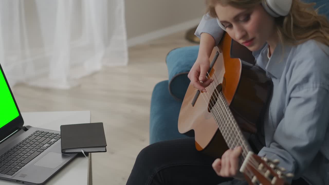 female country musician is playing guitar in room, sitting in front of laptop with green screen, music and hobby