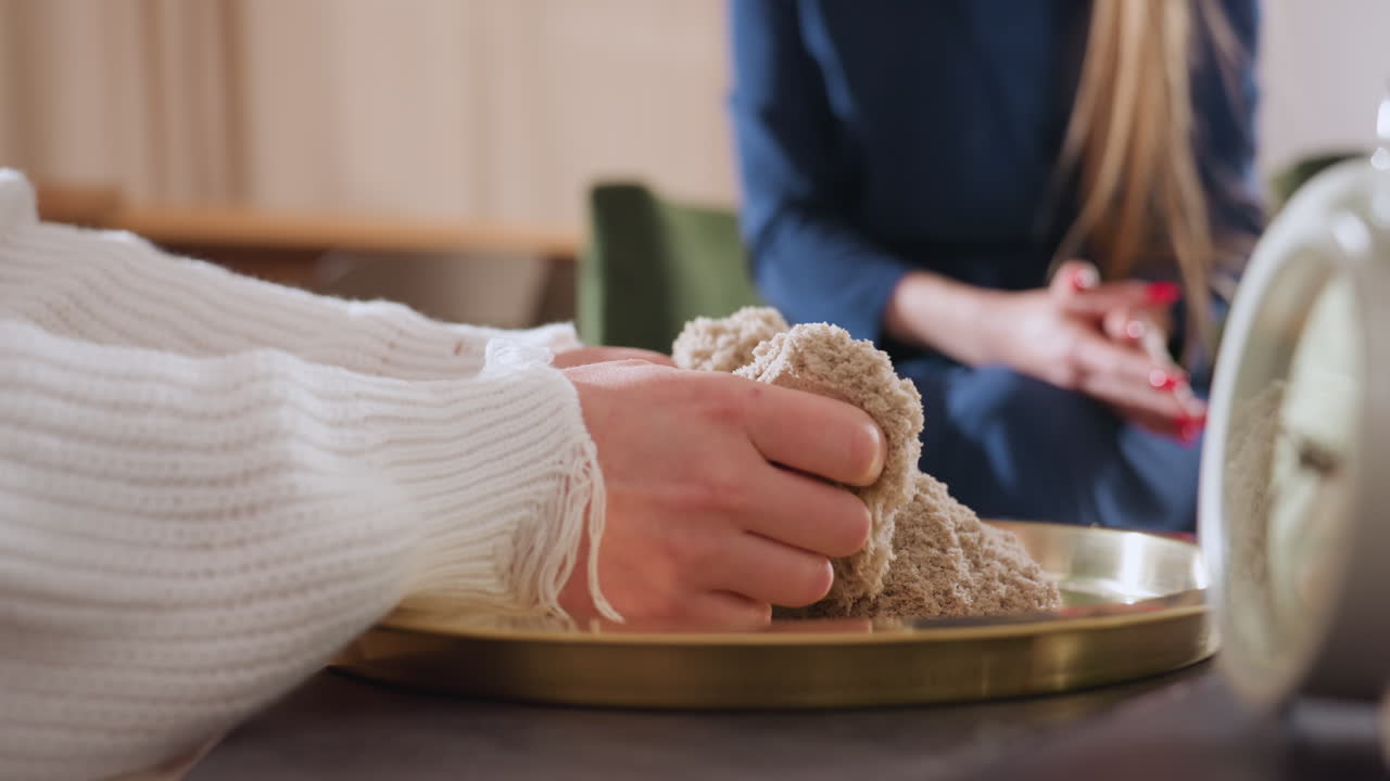 Close up view of woman rubbing hands through soft molded soil on golden tray during therapeutic session, while wellness guide sits nearby in serene room with calm supportive presence