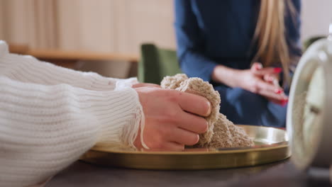 close up view of woman rubbing hands through soft molded soil on golden tray during therapeutic session, while wellness guide sits nearby in serene room with calm supportive presence