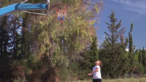 Un-Niño-Caucásico-Juega-Al-Aro-De-Baloncesto,-En-La-Cancha-De-Baloncesto-Del-Callejón,-Hermosos-árboles-En-El-Fondo