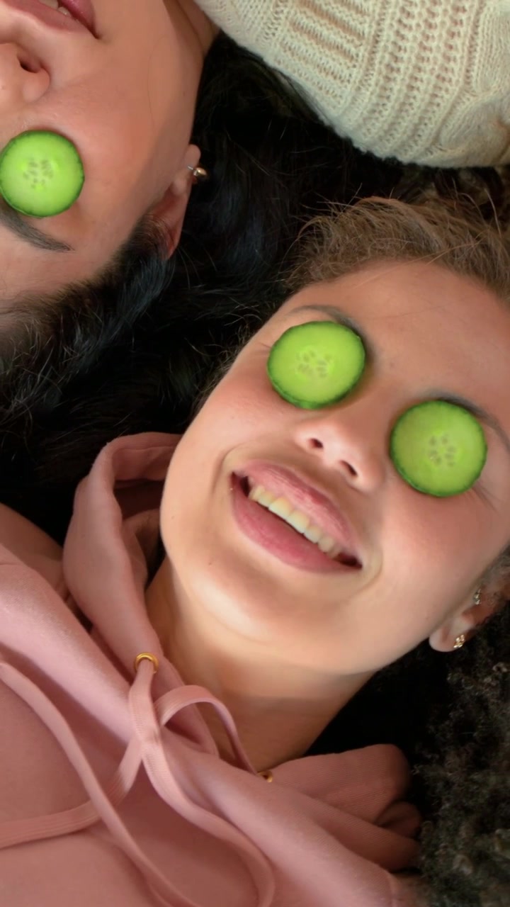 Lying friends wearing pastel striped sweater and pink hoodie keeping cucumbers over eyes on sofa