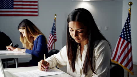 two women writing at desks in a room with american flags, possibly voting or taking an exam