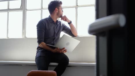 Front-view-of-young-caucasian-businessman-with-laptop-sitting-near-window-in-a-modern-office-4k