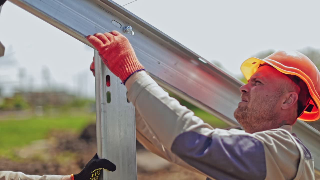 Worker in a uniform and hard hat attaching metal base. Engineer installing metal basis for photovoltaic panel on a solar farm.