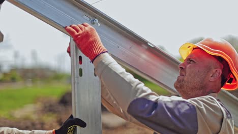 Worker in a uniform and hard hat attaching metal base. Engineer installing metal basis for photovoltaic panel on a solar farm.