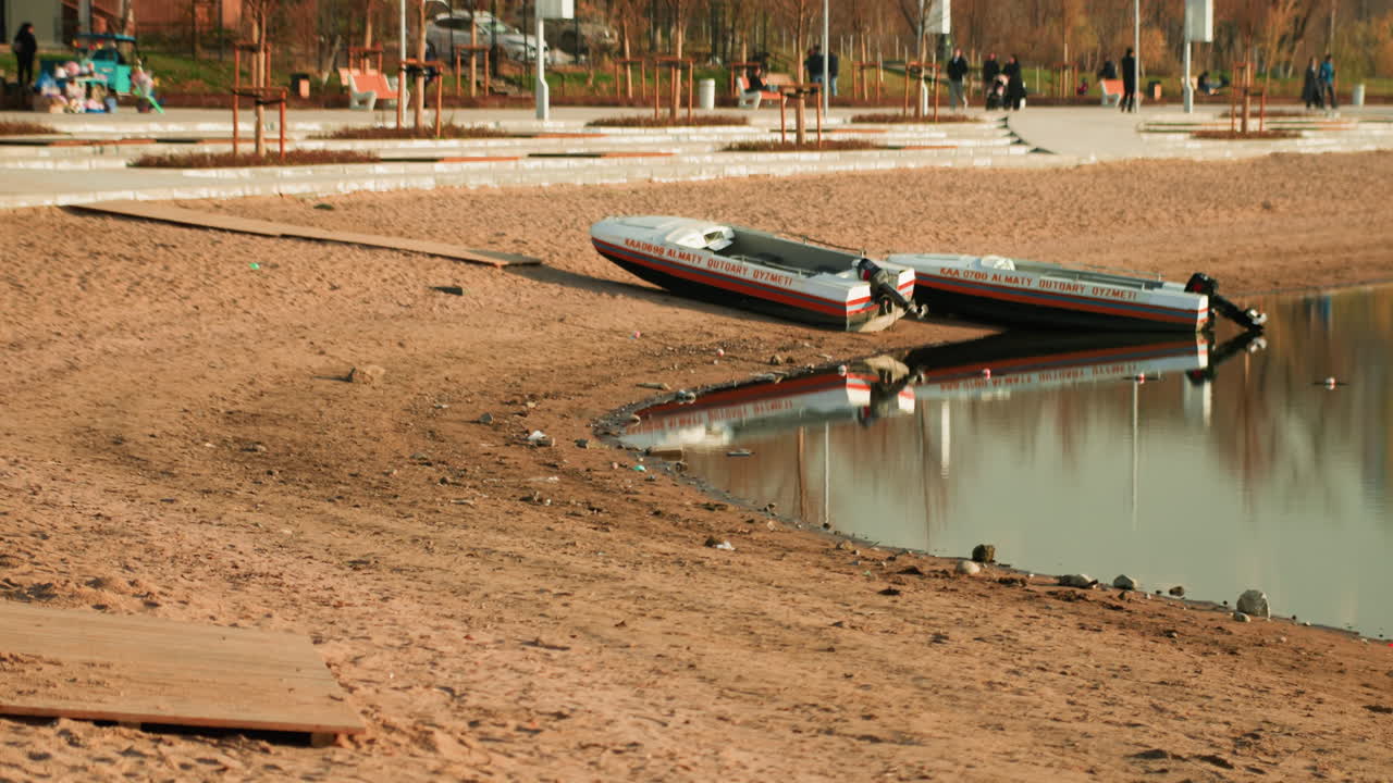 Margem arenosa do lago com barco virado, água calma e reflexiva espelhando o casco, margem lamacenta e areia texturizada, fundo de parque urbano com tons outonais suaves, amarração abandonada evoca solidão