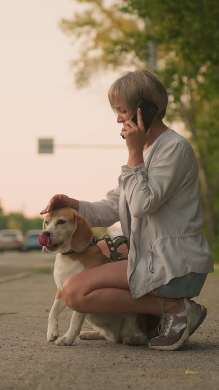 Woman squatting outdoors with brown dog between legs while talking on phone, leash in hand, relaxed scene near rural road with cars passing by, green trees in background