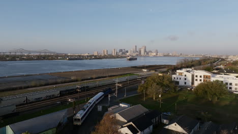 aerial flyover of a passenger train moving towards downtown with a large container ship on the mississippi river with downtown new orleans in the background in the morning