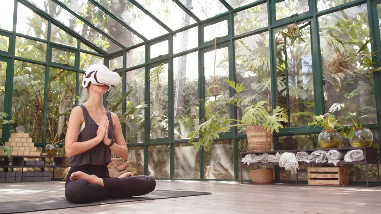 Practicing yoga, woman wearing VR headset in glass house, meditating on mat, copy space