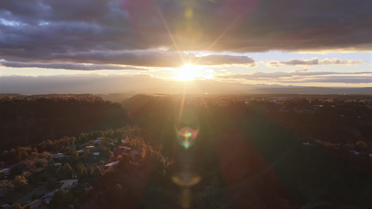 Drone flies straight into the rising sun with warm cinematic lens flares, then smoothly tilts down to reveal a forested mountain valley glowing in golden morning light under dramatic clouds