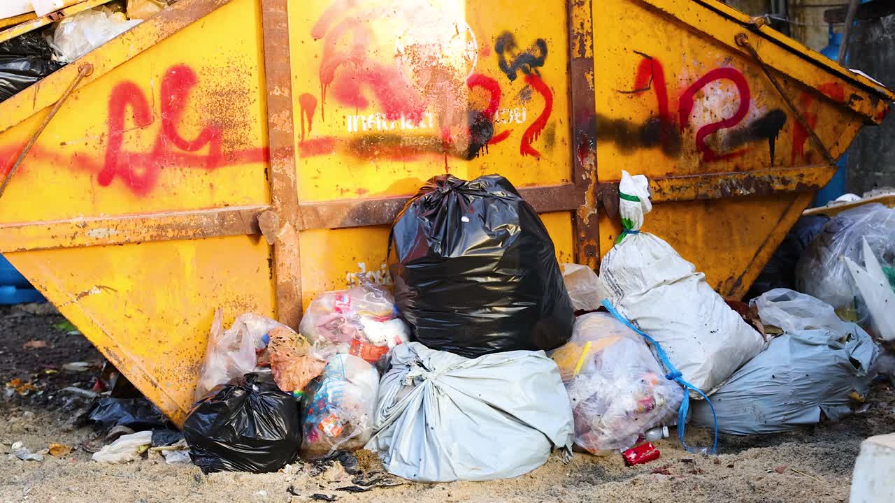 A cluttered dumpster area in Phuket, Thailand, with overflowing trash bags and scattered debris. Bright daylight highlights the urban decay