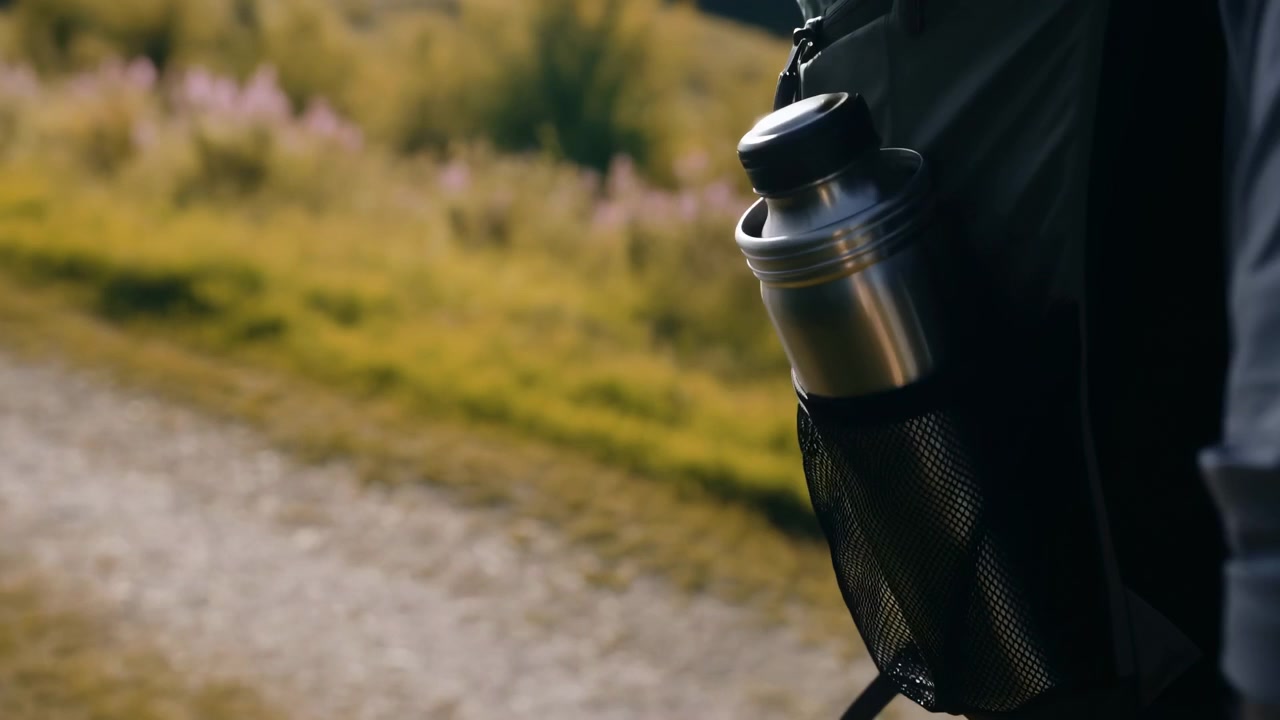 Metal insulated bottle with screw-top lid secured in dark backpack mesh pocket, captured at waist level beside trail scenery with soft sunlight, durable travel and hiking context, with bottle