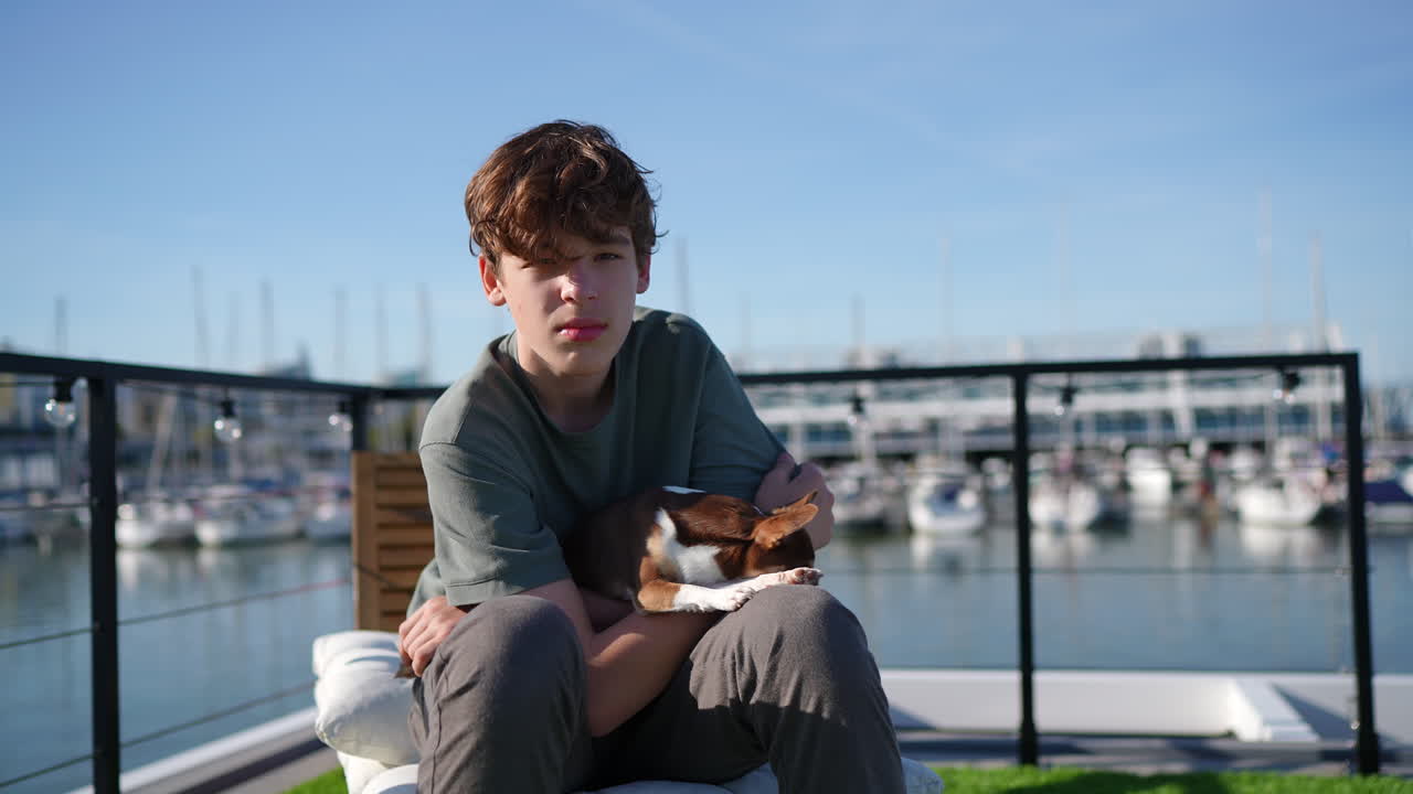 Teenage boy gently holding small chihuahua while sitting on boat at marina, enjoying sunny waterfront moments of companionship and relaxation