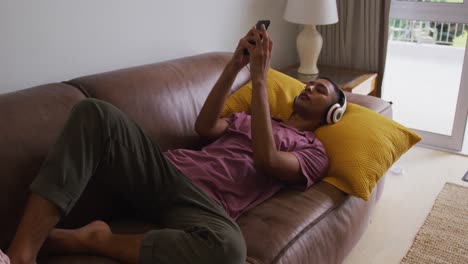 mixed race man wearing headphones lying on sofa using smartphone