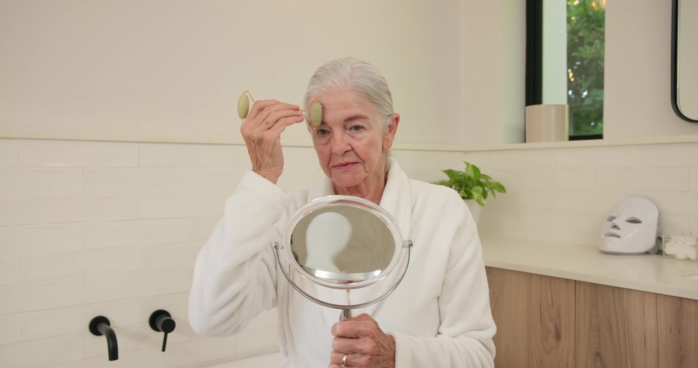 Donna anziana in accappatoio bianco che solleva uno specchio, facendo scorrere un rullo sul viso al lavandino del bagno per la cura della pelle.