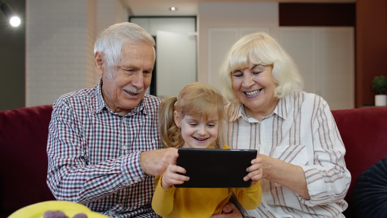 Senior Caucasian couple with cute child girl granddaughter using tablet, watching funny videos