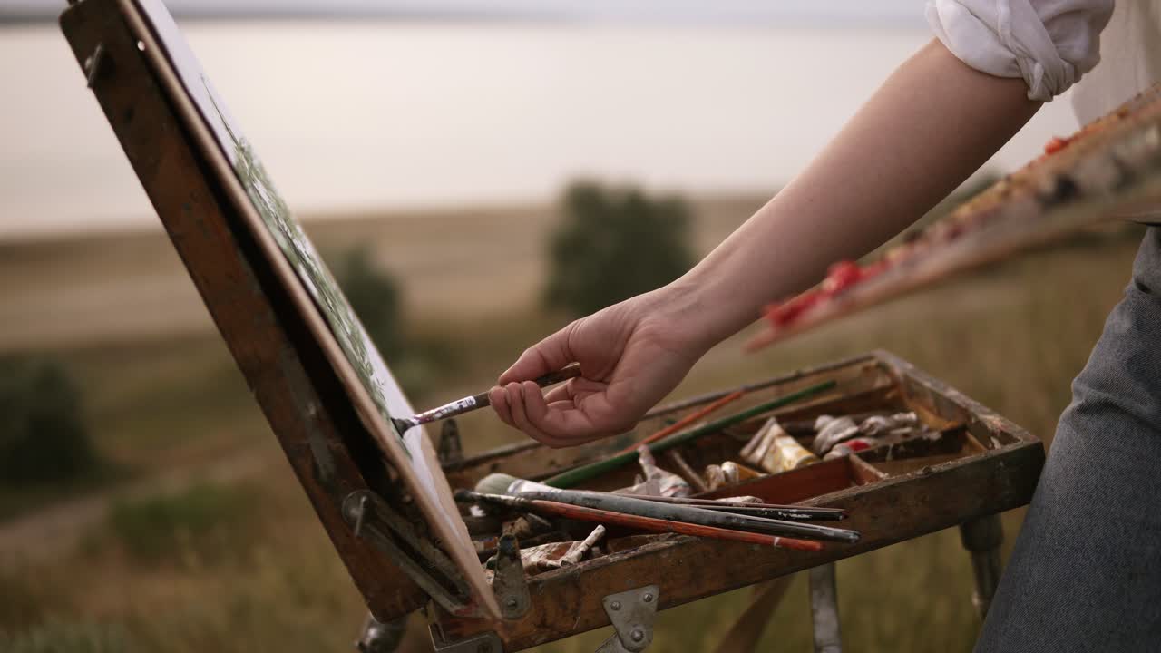 The girl artist in striped shirt mixes the colors on the palette and applies them to the canvas on the easel. Outdoors, wind blowing