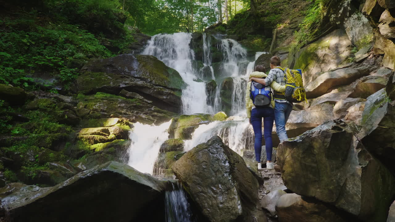 una joven pareja de turistas admira la cascada que se paran en un abrazo la vista trasera 4k 10 bit vid