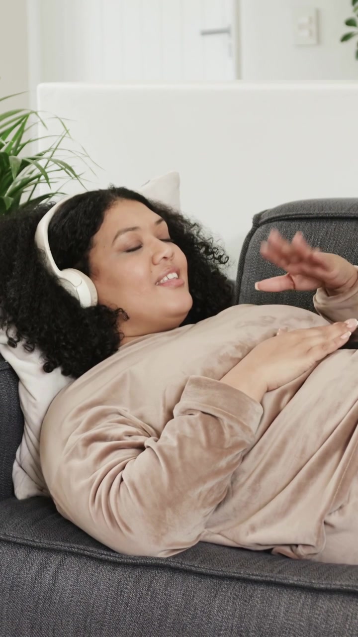 Video verticale: Donna sdraiata sul divano che allunga la mano per prendere le cuffie, toccando lo smartphone per cambiare brano.