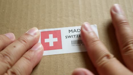 Hände-Bringen-Das-Flaggenetikett-„Made-In-Switzerland“-Auf-Einem-Versandkarton-Mit-Einem-Barcode-In-Premiumqualität-An