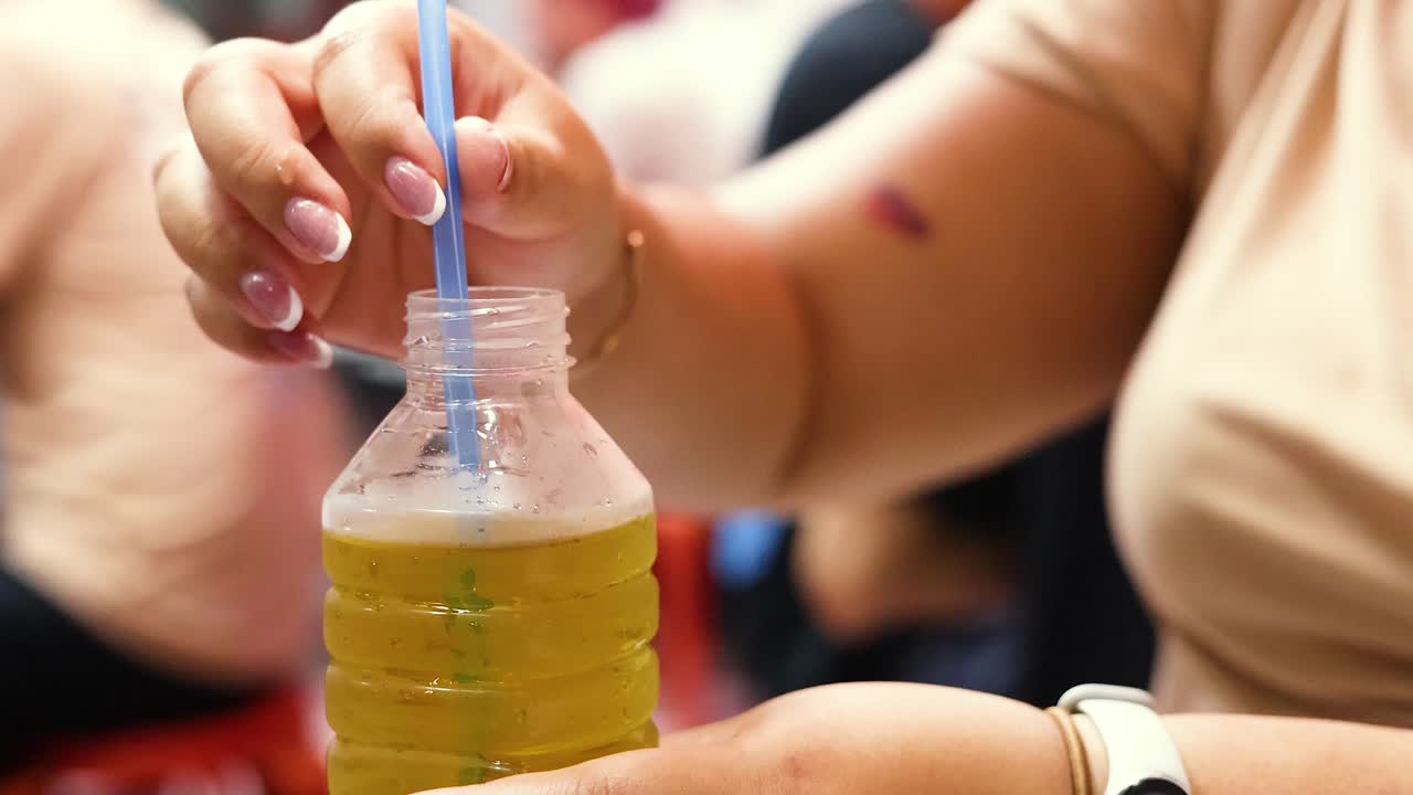 Close-up of woman inserting straw into yellow beverage bottle at busy outdoor market, natural lighting