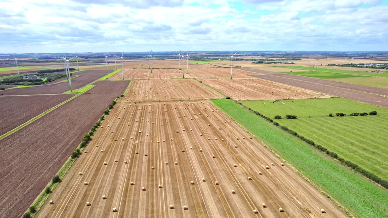 The camera soars above a Lincolnshire farmer's field, revealing an enchanting aerial perspective of wind turbines spinning in the newly harvested landscape, with golden hay bales adding charm