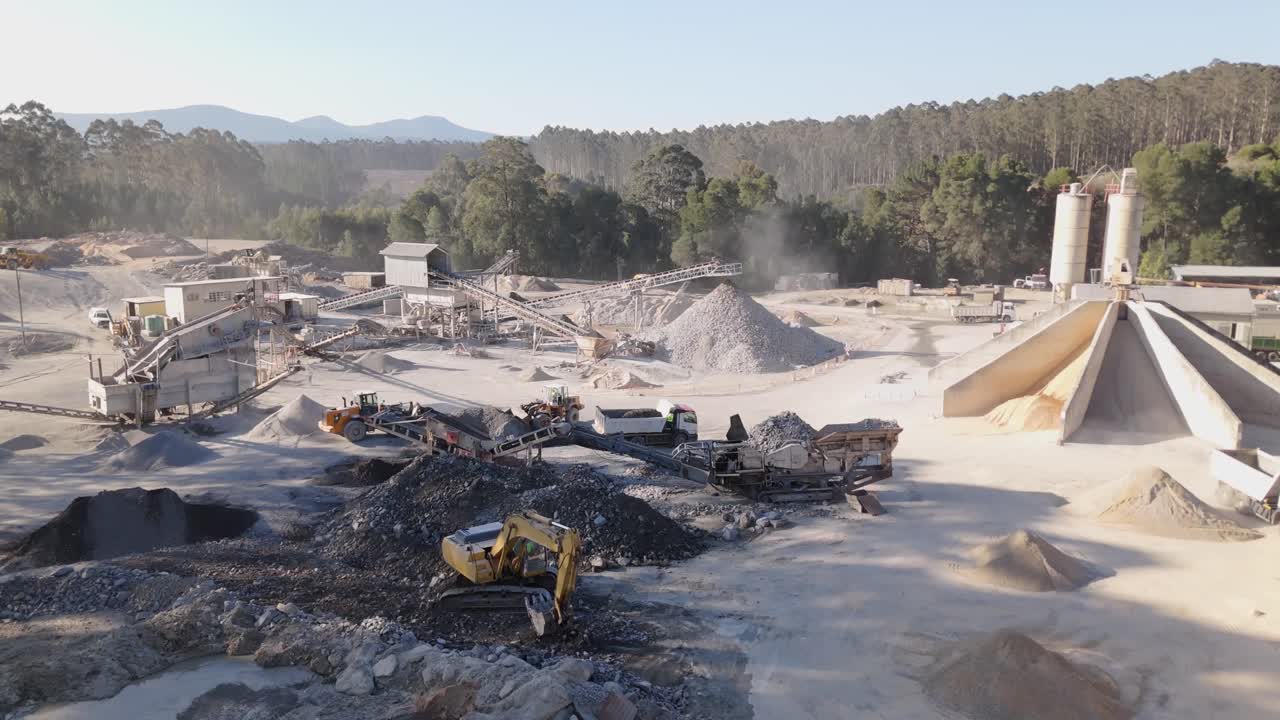 Aerial view of an active stone quarry. Trucks, excavators, and conveyors move rock and sand in dusty conditions