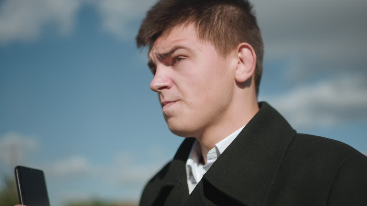 금속 버튼을 은 검은 코트를 입은 사업가, 은 파란 하늘 아래 밖에서 전화 통화를 하고, 흐릿한 초록색과 현대적인 도시 배경으로 상하고 있습니다.