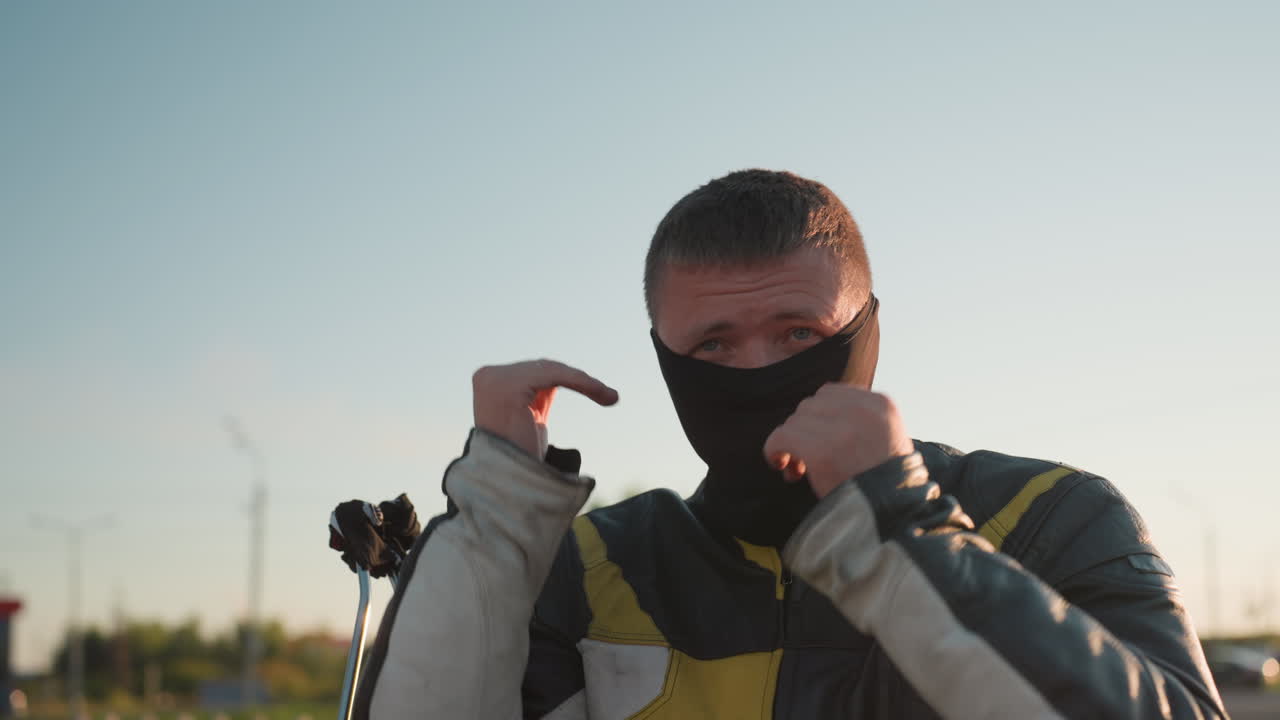 Tough man in leather jacket adjusts collar while standing outdoors under warm sunlight glow, preparing for ride with determined expression as sun flare brightens background atmosphere