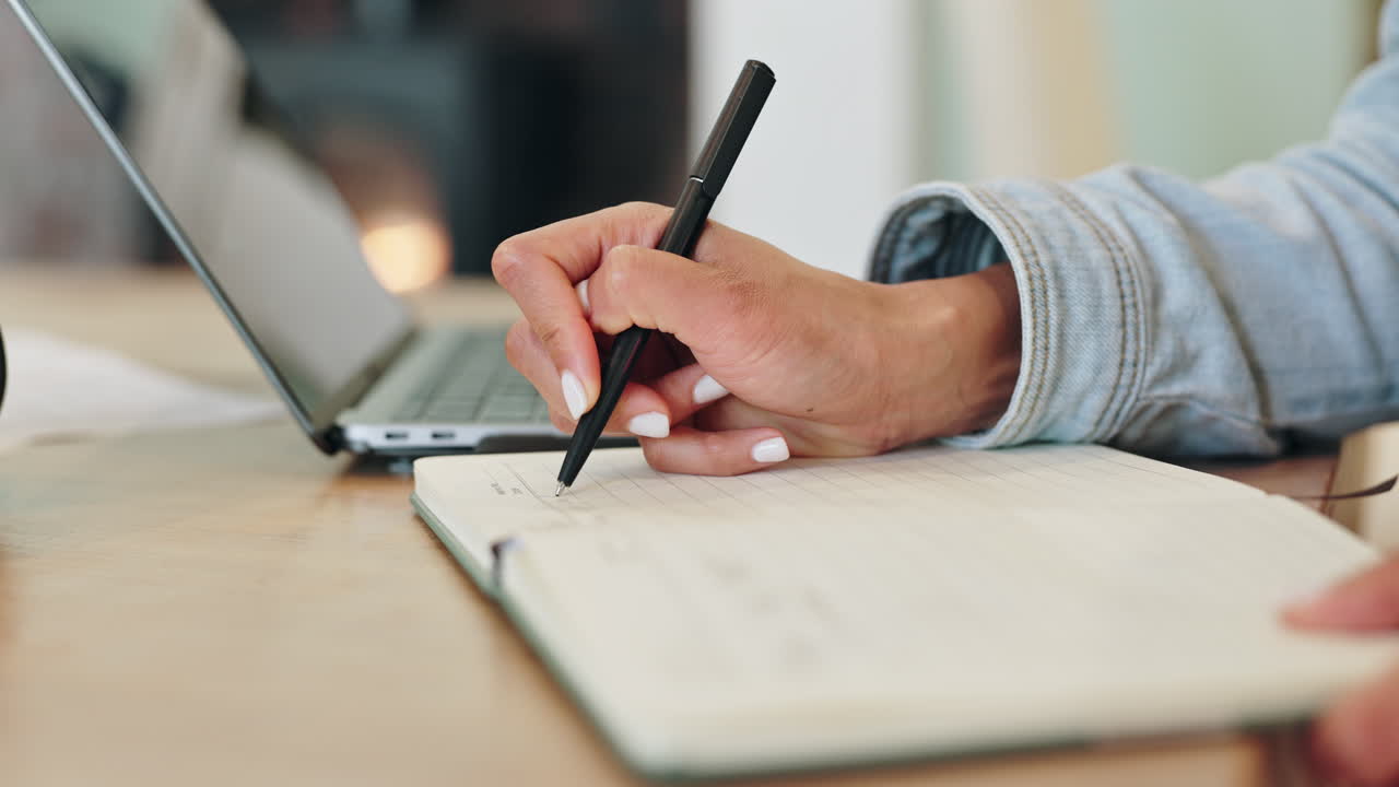 Person writing in a notebook with a pen next to a laptop