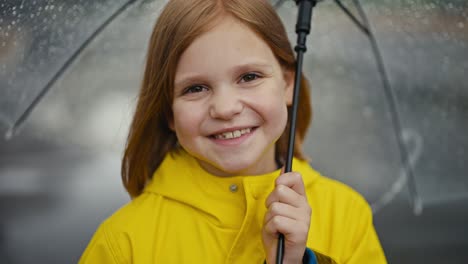 portrait of a happy blonde teenage girl in a yellow jacket holding a transparent umbrella with water drops in the park while walking after the rain